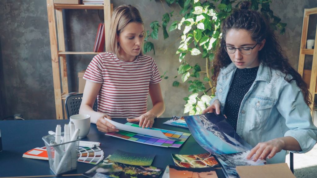 Two women collaborating on a design project, reviewing printed materials and color swatches.